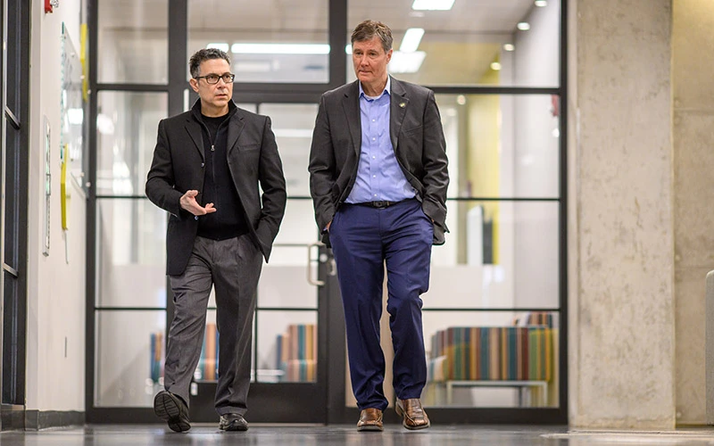 Two men in business attire walk and talk in a hallway with glass-walled rooms.  The man on the left gestures with his hand as he speaks.