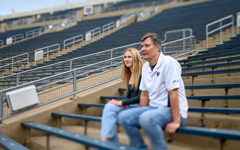 A man and a young woman sit in the empty bleachers of Notre Dame Stadium. The man wears a white Notre Dame polo shirt and jeans.  The woman wears a dark green shirt and light-wash jeans.  They are looking towards the field.