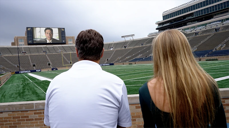 Two people, seen from the back, watch a large video screen inside Notre Dame Stadium. The screen displays a man in a suit.