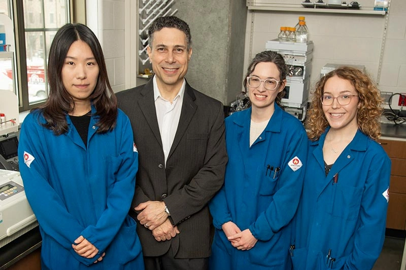 Four individuals, three in blue lab coats and one in a suit, stand together smiling in a laboratory setting.