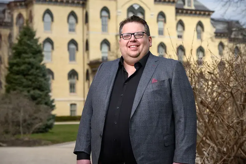 A man in a gray suit and black shirt smiles in front of a light yellow building on the University of Notre Dame campus.