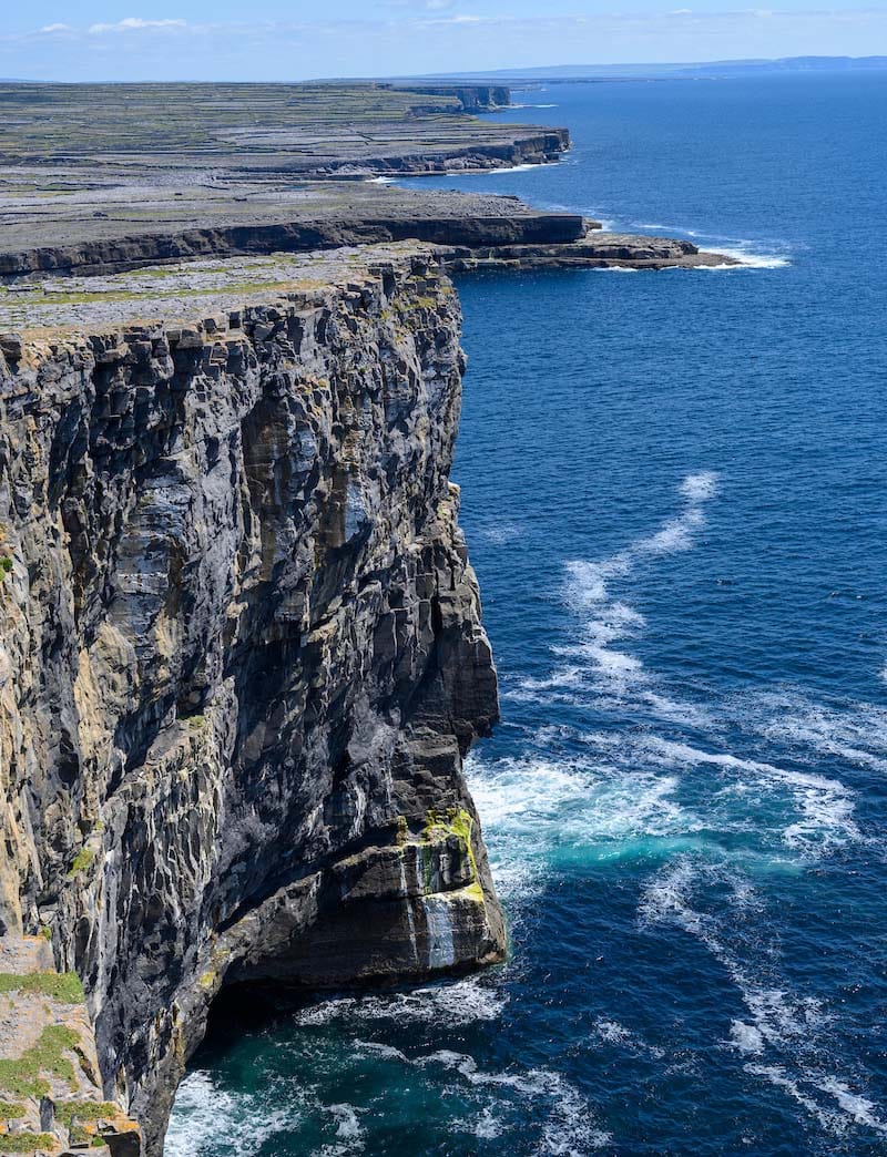 Gray stone cliff high above blue crashing ocean below.