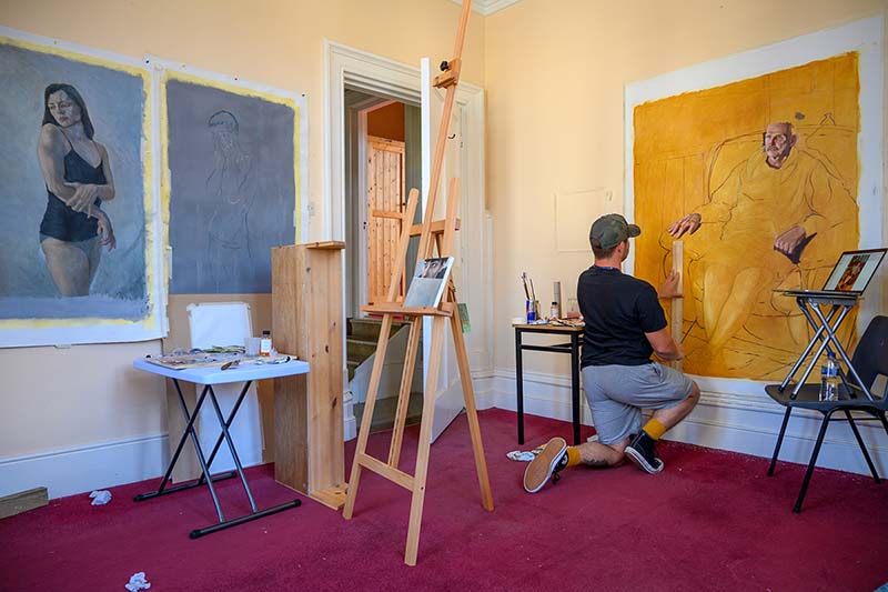 A man paints a gold portrait of a man. A portrait of a lady hangs on the wall behind him.