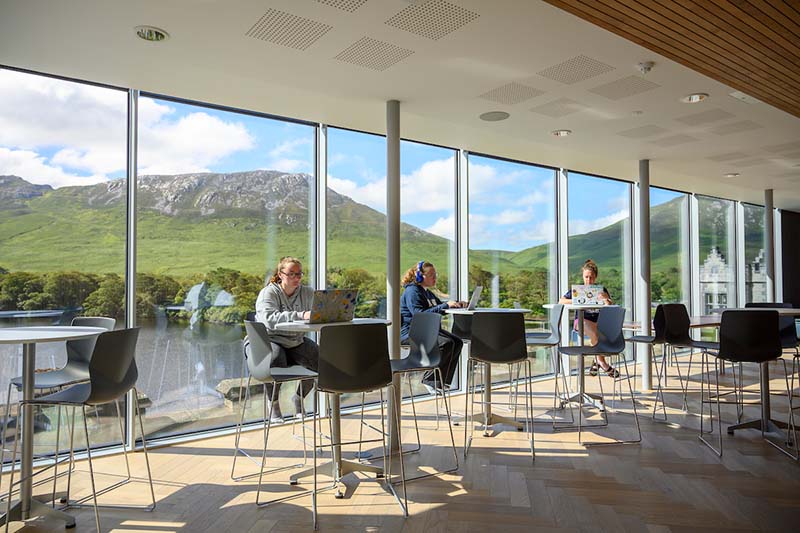 Students study at tables with a wall of windows behind them providing a view of green mountains and the lake.