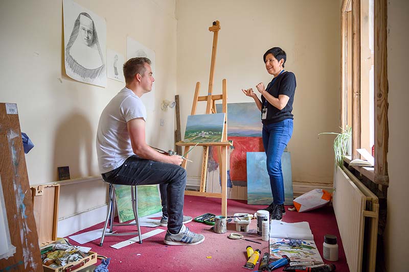 A man sits in front of a painting with an easel in his hand. A woman speaks to him.
