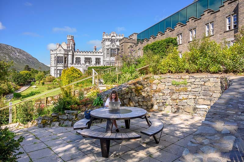 A girl sits at a table with a laptop outside the buildings.