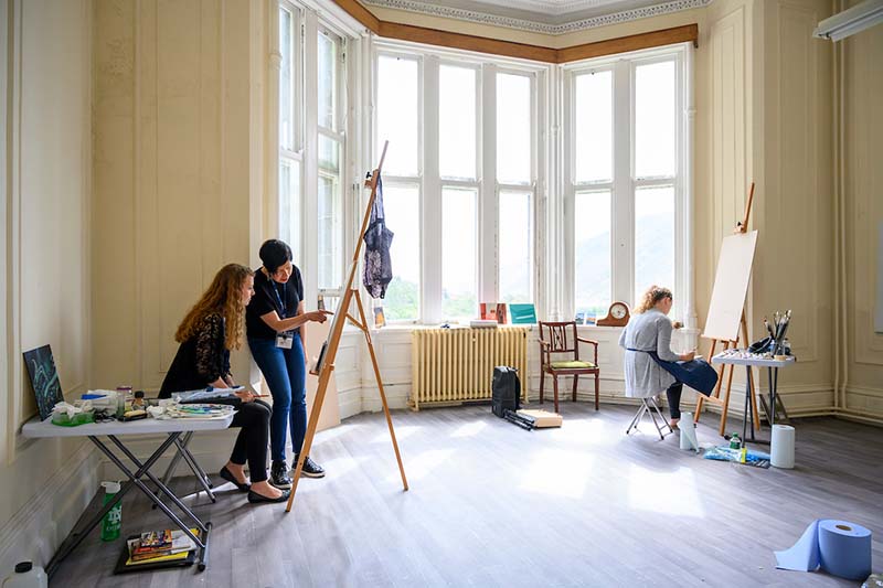 Two students sit before easels, while a woman helps one of them. Large windows let in lots of sunlight.