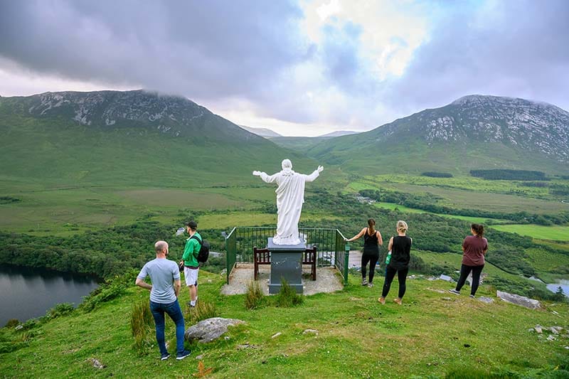 Students stand next to an arms-outstretched statue of Mary, overlooking green hills and lakes.