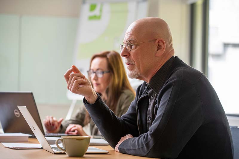 A man and a woman sit at a table with laptops in front of them. The man is talking.