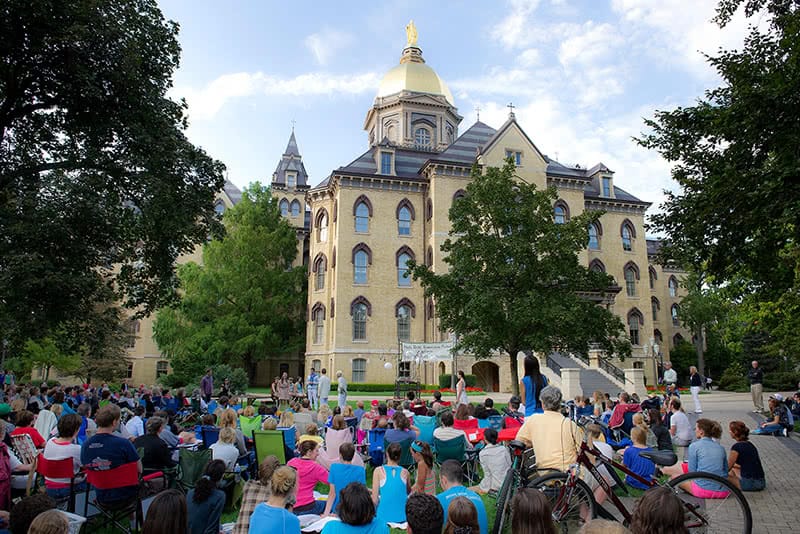 A large group of people seated on chairs and the ground in front of the Main Building