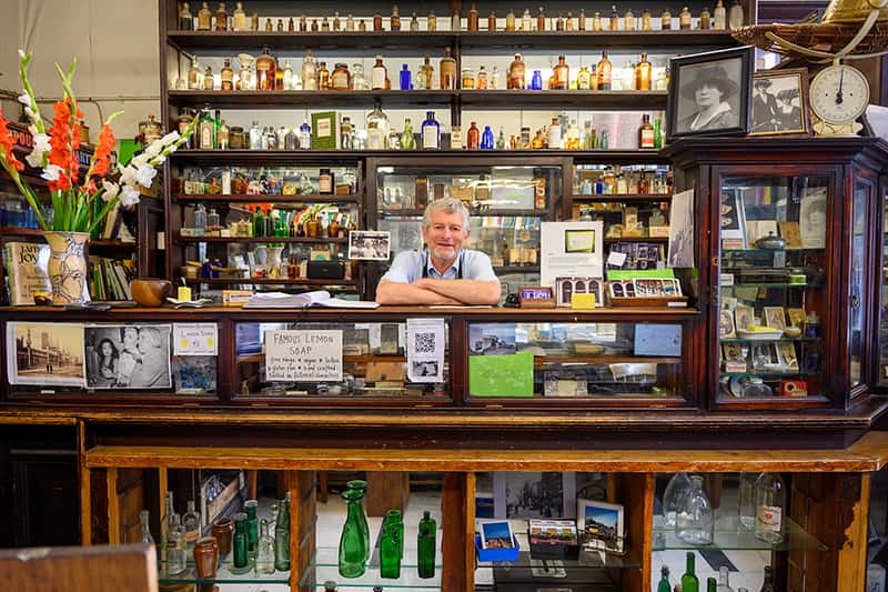 A man stands behind a counter surrounded by glass bottles.