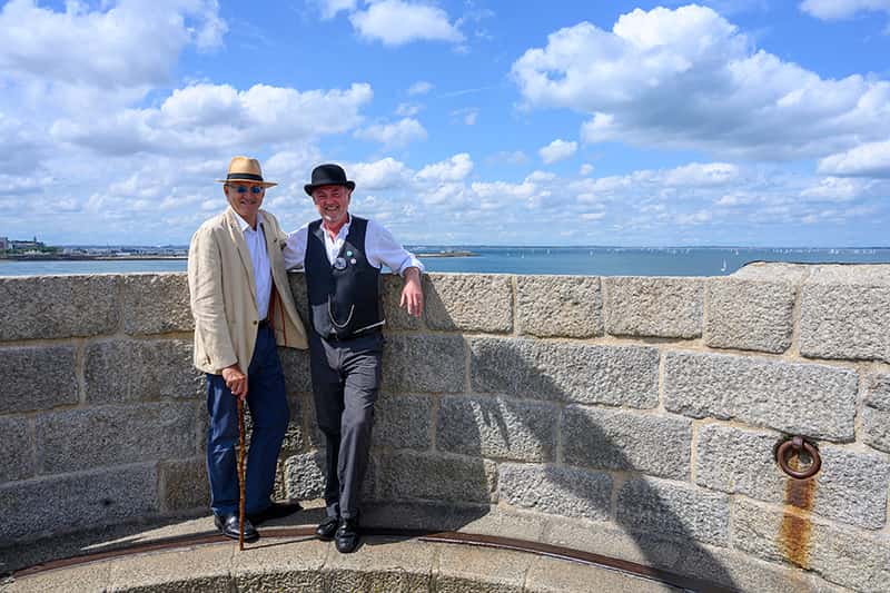 Two men stand at the top of a tower, against a wall. The blue sky, clouds, and the ocean is in the background.