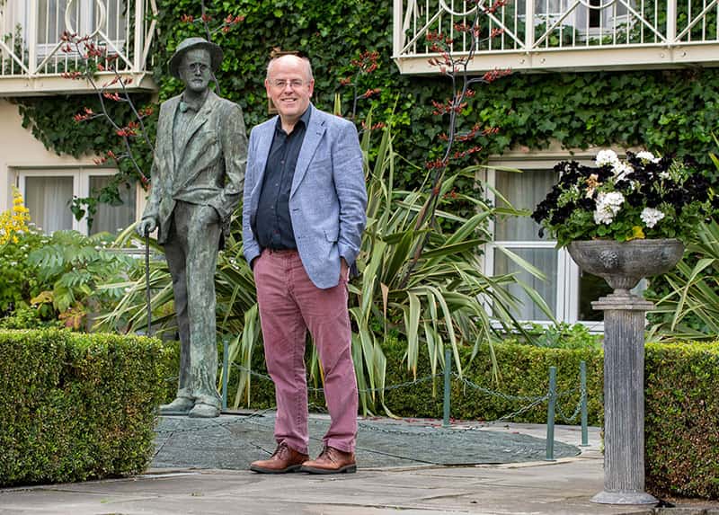 With his hands in his pocket, a man stands in a garden next to a statue of James Joyce.