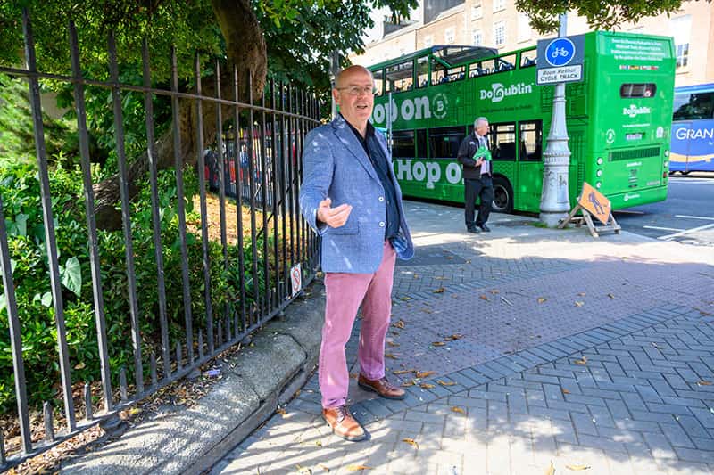 A man stands on a street corner telling a story. There's a green double decker bus and people walking in the background.