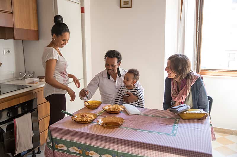 Three adults and a child sit and stand around a table with four bowls full of pasta.