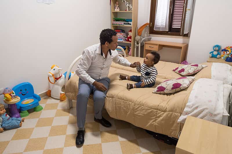 A father sits on a bed talking to his son.