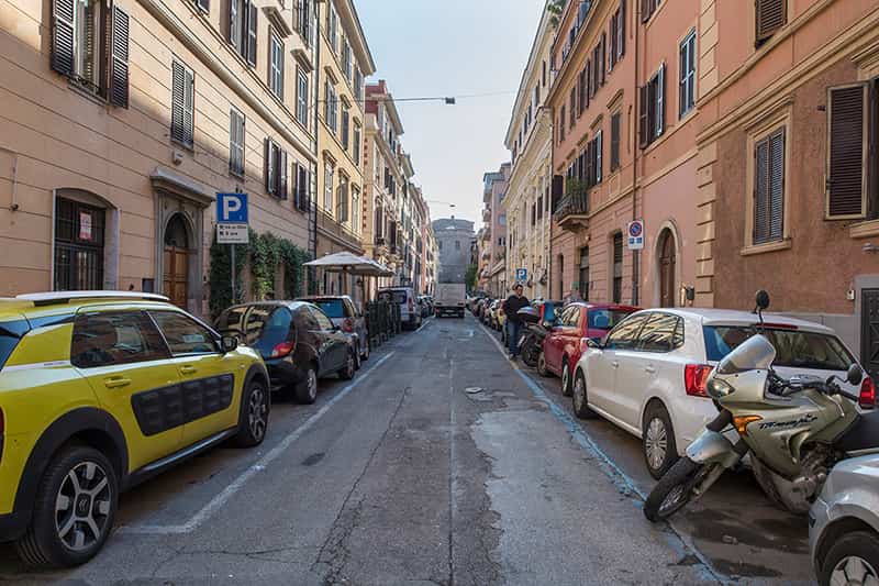 A narrow road, buildings and parked cars line both sides.