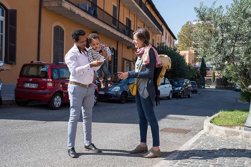 A woman speaks to a man who is holding a child outside of an apartment building.