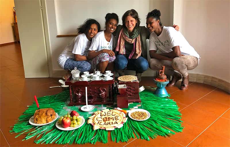 Four people pose for a photo as they squat down next to food and drinks that are set on the floor.