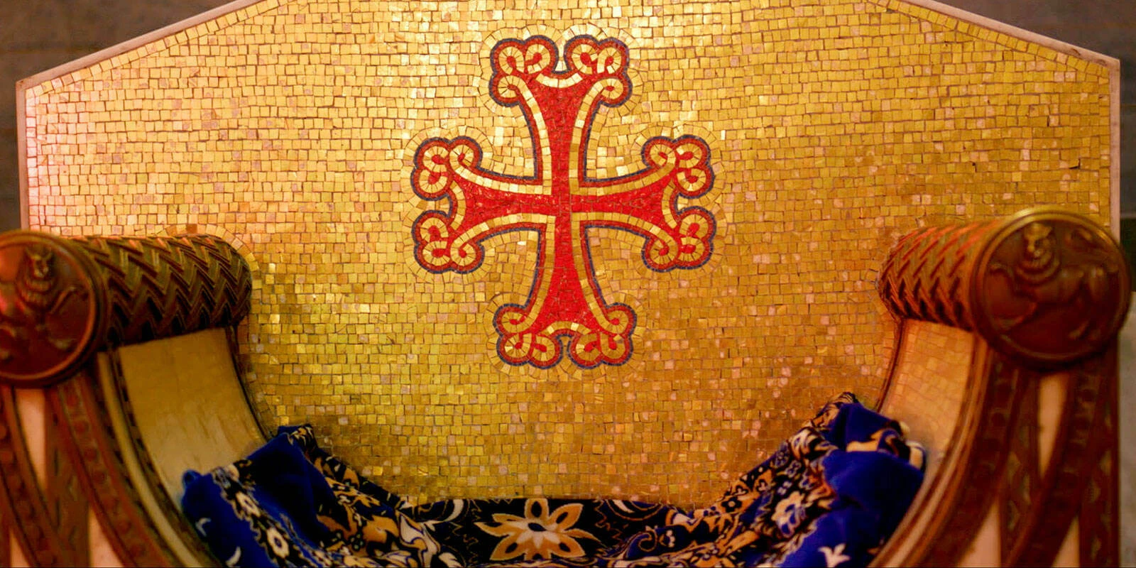 A golden mosaic wall with a large red cross, with ornate cushioned pews in the foreground covered by a blanket.