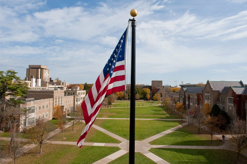 American flag waving on a sunny autumn day on the University of Notre Dame campus.  The quad, crisscrossed by walkways, and several campus buildings are visible in the background.
