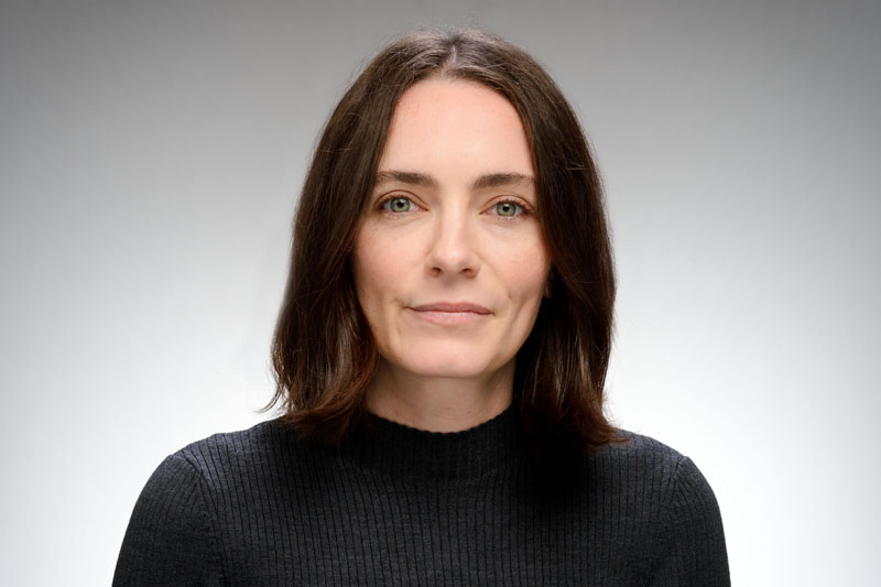 Headshot of a woman with shoulder-length brown hair and green eyes. She wears a dark gray ribbed turtleneck sweater and offers a slight, closed-lip smile.