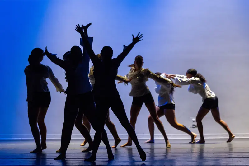 Dancers on a stage in silhouette with a blue background, their arms and legs extended in various poses.