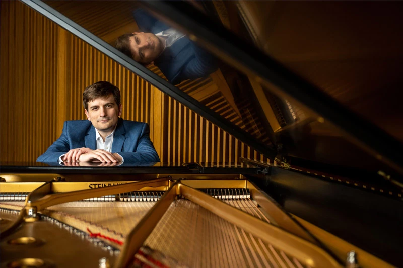 A man in a blue suit and white shirt sits at a grand piano, his hands resting on the keys. A reflection of his face is visible on the piano's polished lid.