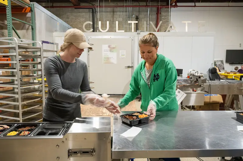 Two women, wearing gloves, package meals in a commercial kitchen with &rdquo;CULTIVATE&ldquo; on the wall and food trays in the background.