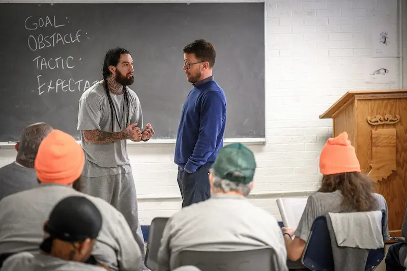 Two men stand facing each other in front of a blackboard addressing a seated audience.
