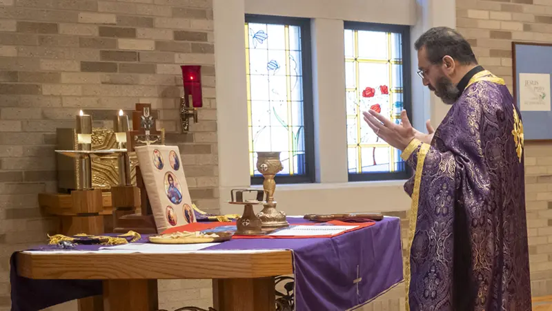 A priest in a rich purple vestment stands before an altar with lit candles, reciting prayers with his hands raised near stained-glass windows.