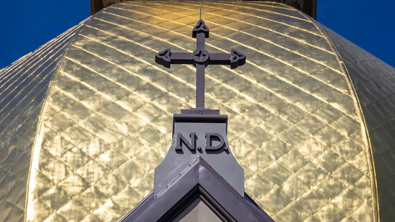 A close-up of the top of the University of Notre Dame's Golden Dome, featuring a dark cross with Celtic knot details above the letters 'N.D.'