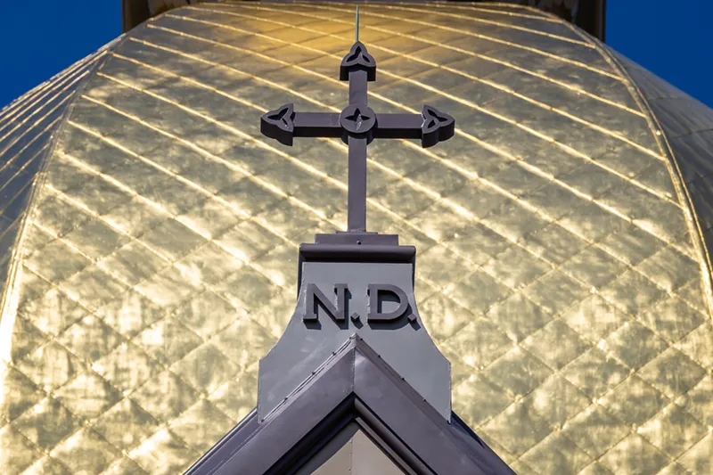 A close-up of the top of the University of Notre Dame's Golden Dome, featuring a dark cross with Celtic knot details above the letters 'N.D.'