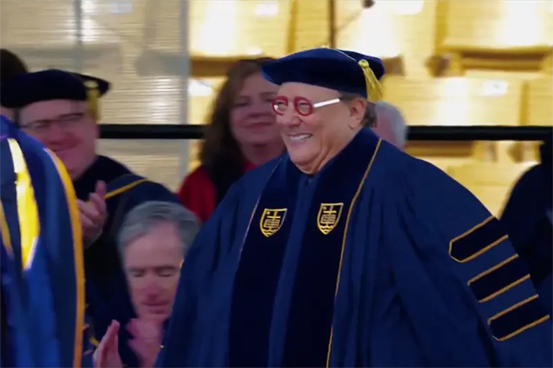 A smiling man wearing dark blue academic regalia with gold accents and distinctive red-rimmed glasses walks past clapping attendees at a commencement ceremony.
