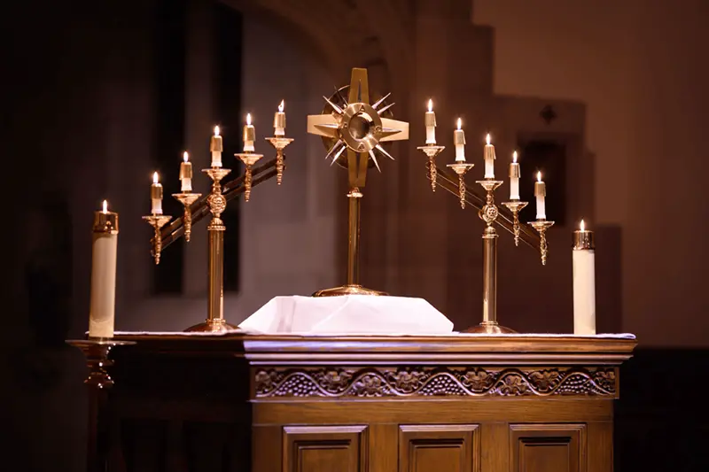 A richly carved wooden altar displays a golden monstrance with spiky rays, surrounded by numerous lit taper candles and two large pillar candles.