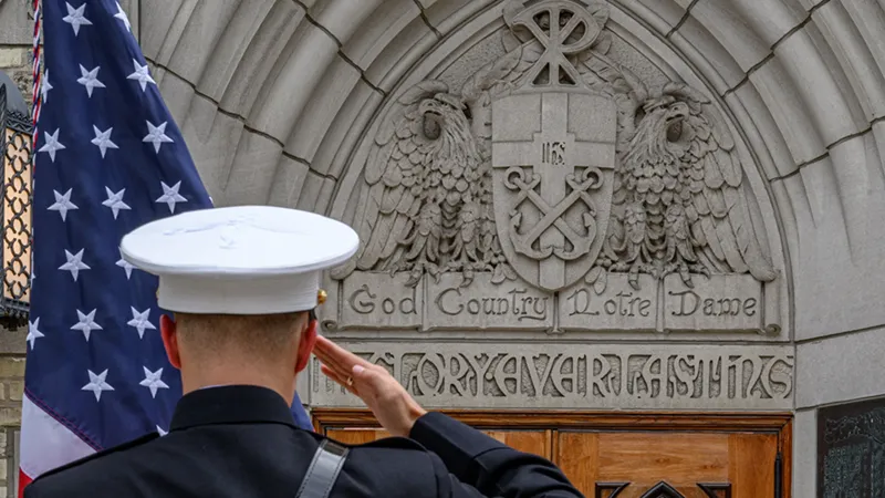 A service member in uniform renders a salute next to the U.S. flag before a stone archway bearing the University of Notre Dame seal and the motto 'God Country Notre Dame.'