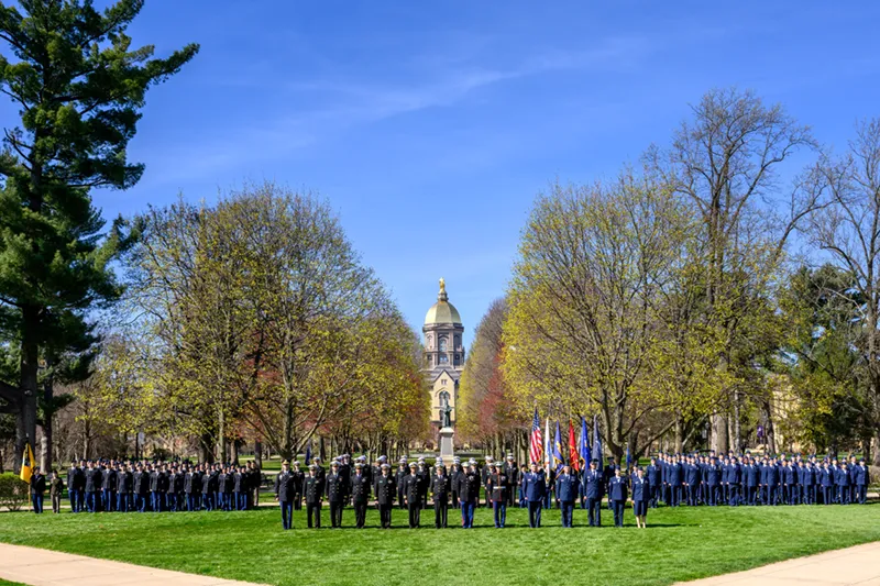 A large formation of ROTC cadets stands on the University of Notre Dame lawn, with the Golden Dome visible between two rows of trees.
