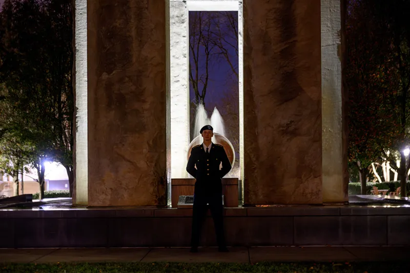 A lone person in a dark military uniform and beret stands at attention in front of a tall, illuminated stone fountain at night.