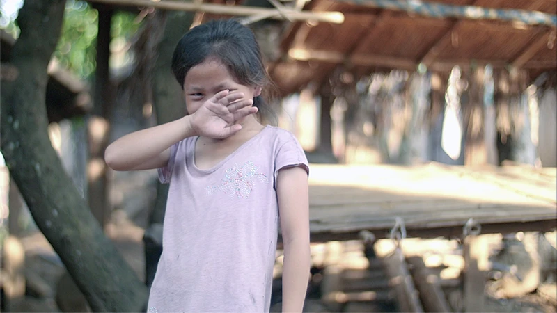 A young girl, wearing a light purple shirt, stands outside and smiles shyly while raising her hand to her face.