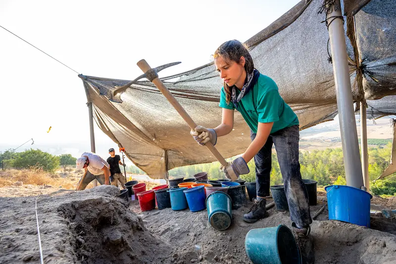 A female archaeologist wearing a teal shirt, gloves, and a bandana around her neck uses a pickaxe at an excavation site with colorful buckets, while other archaeologists work nearby under a tarp.