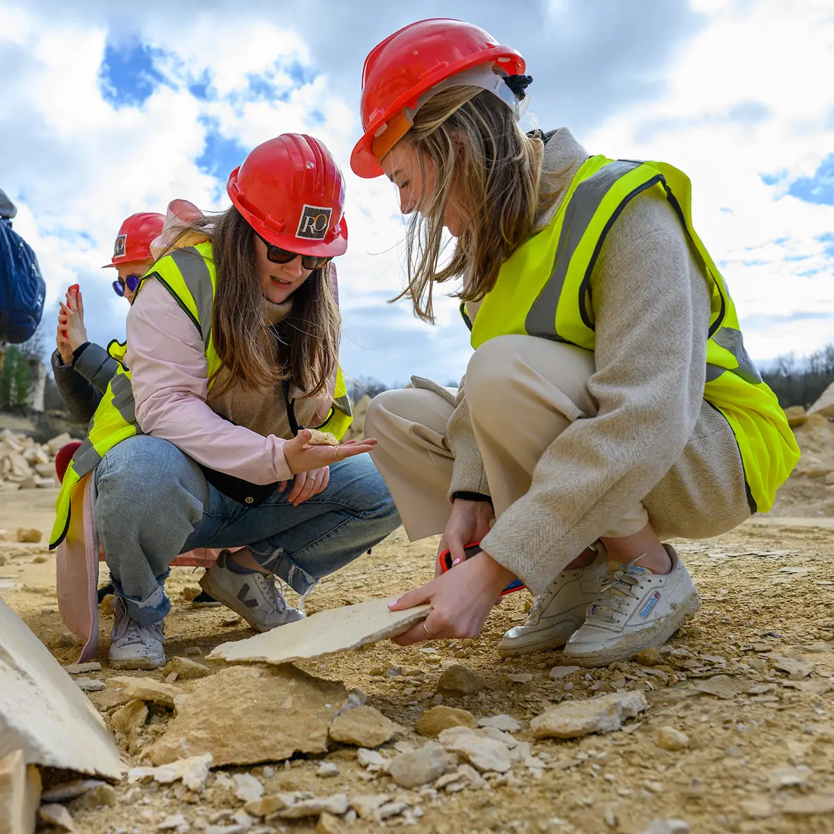Two female construction workers in hard hats and safety vests examine a piece of rock at a work site.