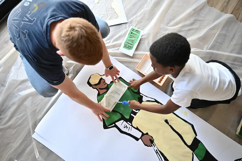 A man and a child are on the floor, collaborating on painting a large stencil of a baseball player.