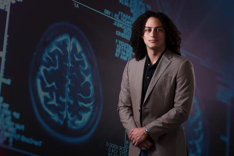 A young man with curly hair in a brown suit jacket stands in front of a blue screen displaying brain scans.