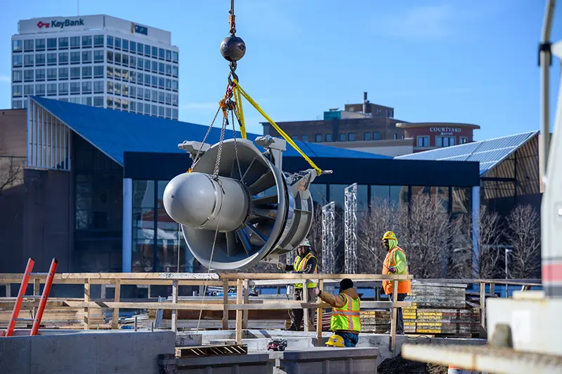 A construction worker in a hard hat and safety vest observes a large, metal turbine being lifted by a crane under a bright, sunny sky.