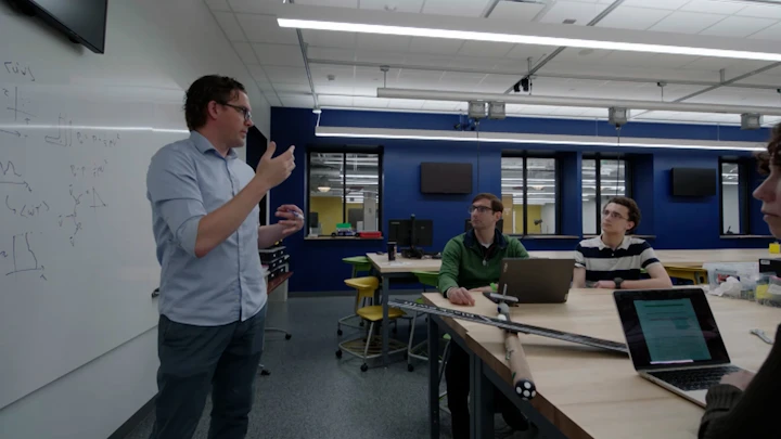 David Richter stands in front of a whiteboard speaking to a few students in a classroom going over classroom material.
