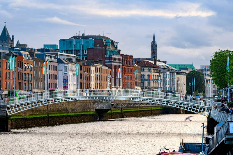 The Ha'penny Bridge crosses the River Liffey in Dublin, with colorful buildings and Irish flags lining the north bank.