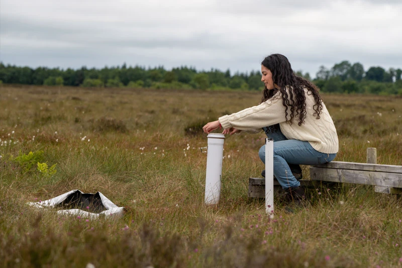 A young woman in a cream-colored sweater kneels on a wooden plank, working with scientific equipment in a grassy, open field.
