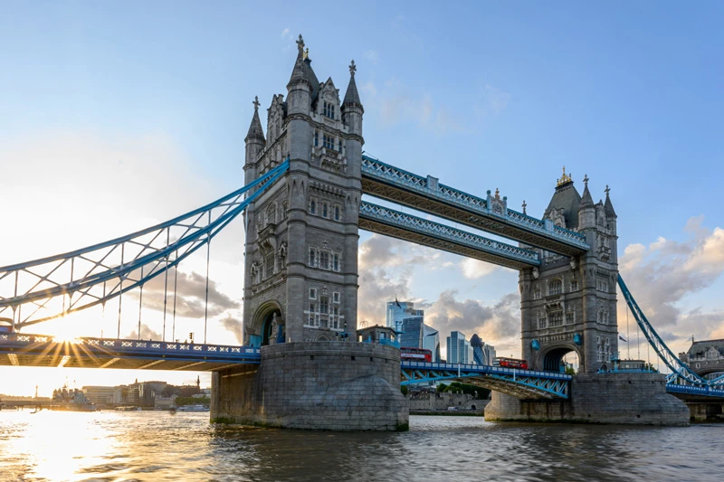 The iconic Tower Bridge in London is lit by the setting sun, with a double-decker bus on the lower deck and city skyscrapers visible in the background.