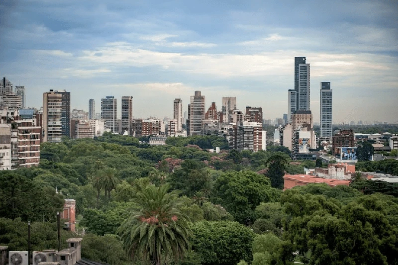 A panoramic view of a city skyline with modern skyscrapers rising above a lush, green canopy of trees.