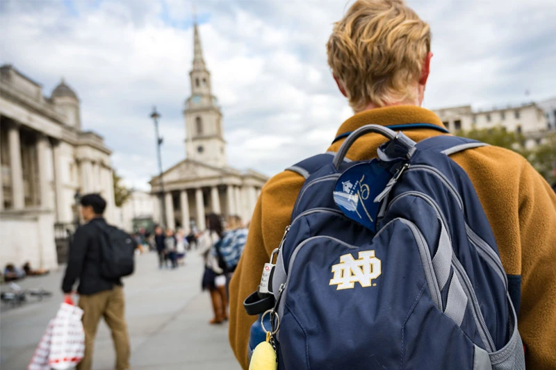 A person wearing a mustard-colored fleece and a blue Notre Dame backpack walks toward a busy plaza with a classical-style church in the background.